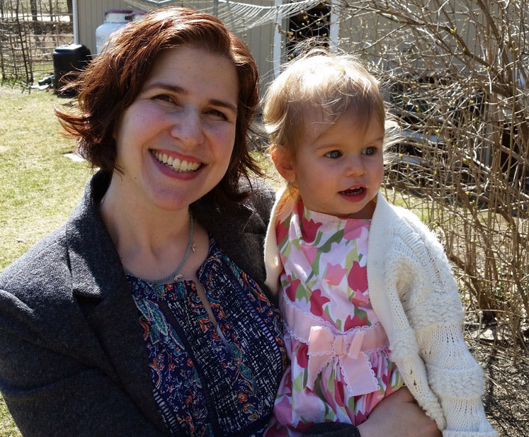 A woman with short brown hair and a broad smile holds a young child dressed in a pink floral dress and a white cardigan. They are standing outdoors on a sunny day with bare bushes and a shed in the background. Both appear happy and content.