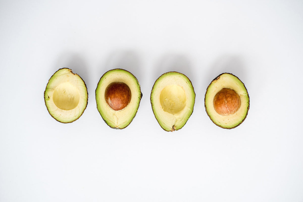 Four halved avocados are displayed in a row on a white background. Two avocados have their seeds intact, while the other two are without seeds. The avocados are cut lengthwise, showing the green flesh and dark skin.