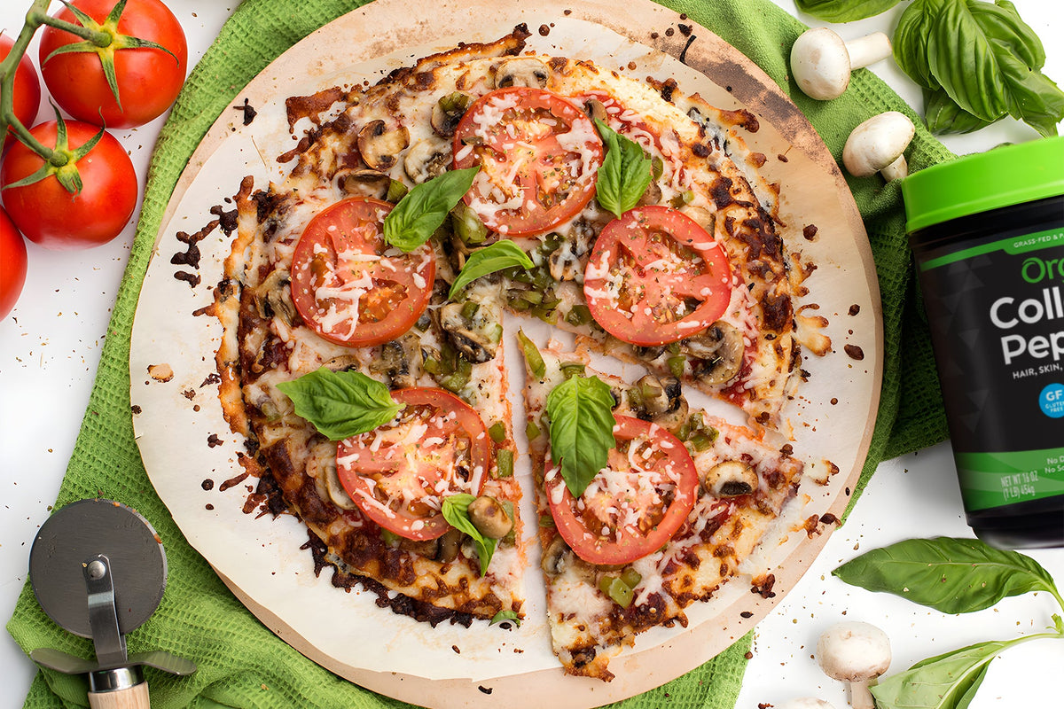 A freshly baked pizza topped with sliced tomatoes, basil leaves, mushrooms, and cheese sits on a pizza stone over a green cloth. A pizza cutter, fresh tomatoes, mushrooms, basil leaves, and a container of collagen peptides are arranged around the scene.