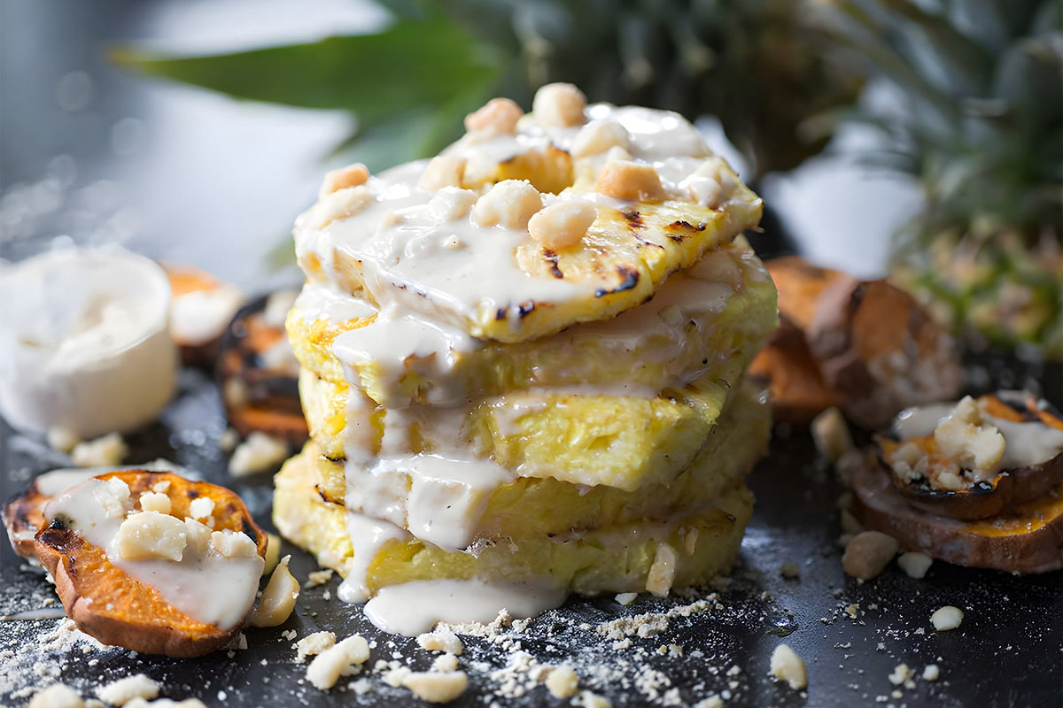 A stack of grilled pineapple slices topped with a creamy white sauce and sprinkled with crushed nuts sits on a black plate. Surrounding the stack are additional grilled slices of sweet potato and some white cheese, with a blurred green leafy background.