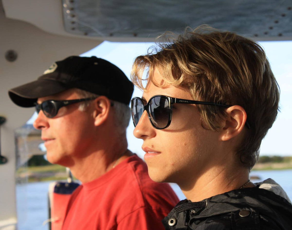 Two individuals in a boat wearing sunglasses, looking forward. Water and trees are in the background.