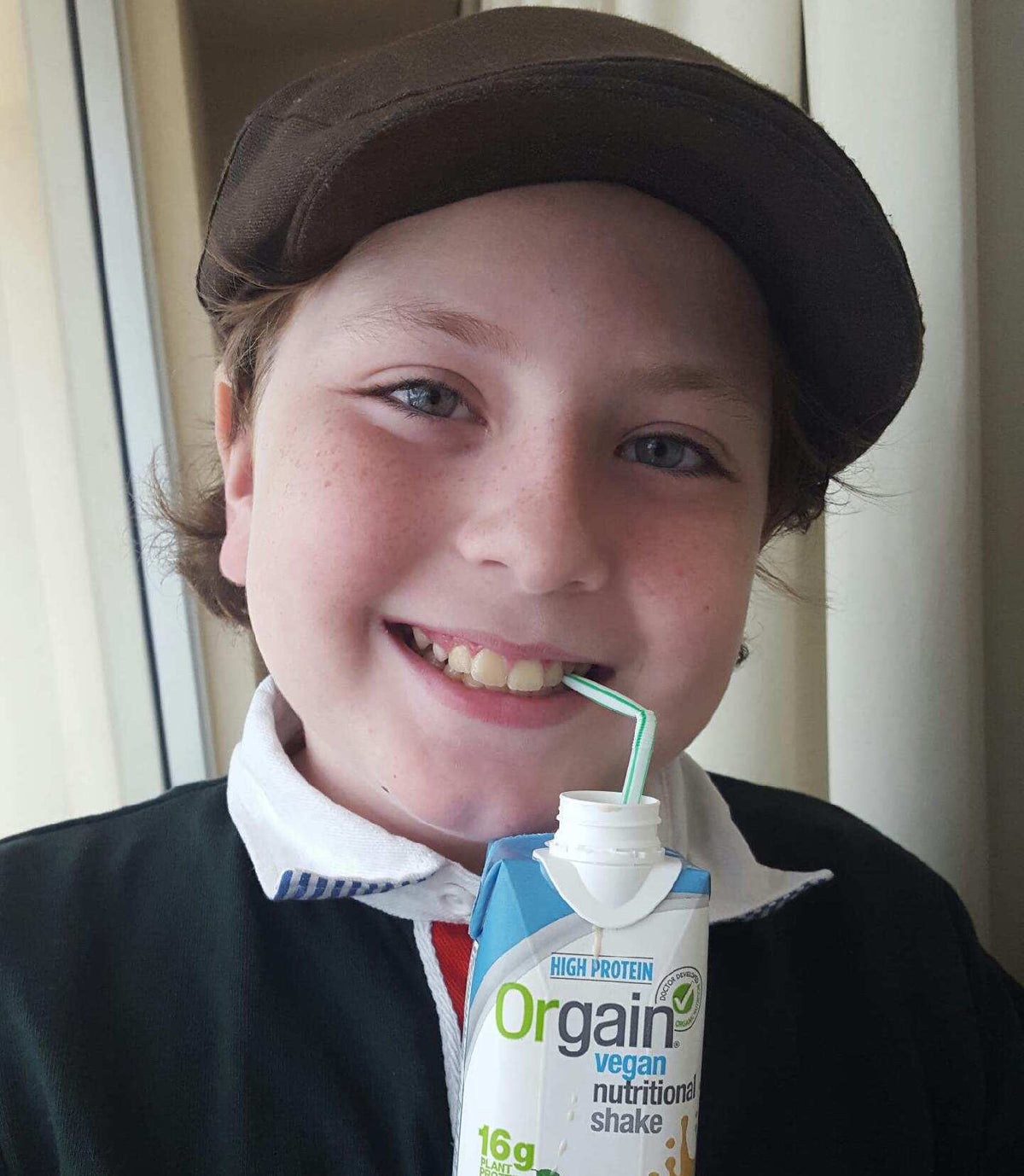 A smiling child wearing a brown cap and dark jacket drinks from a straw inserted into a carton of Orgain vegan nutritional shake. The background features light-colored curtains. The child has light skin and short, light brown hair.