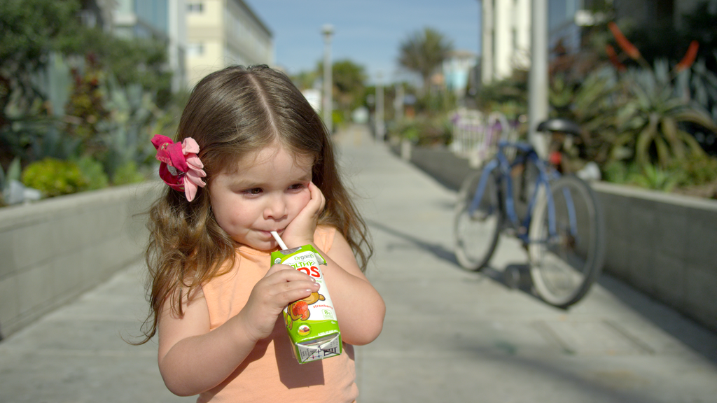 A young girl with long brown hair and a pink flower hair clip is sipping from a juice box. She stands on a sunny sidewalk, with a bicycle and greenery visible in the blurred background. The girl is wearing a peach-colored shirt and looks thoughtful.