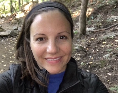 A woman with a headband and a smile takes a selfie in a forested area. She is wearing a dark jacket and has trees and a dirt path in the background.