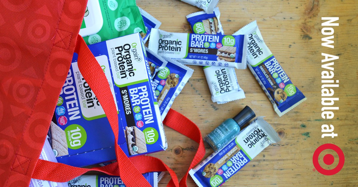 A collection of protein bars and small wellness products are partially spilling out from a red Target shopping bag on a wooden surface. Text on the right side reads, ‚ÄúNow Available at‚Äù followed by the Target logo.