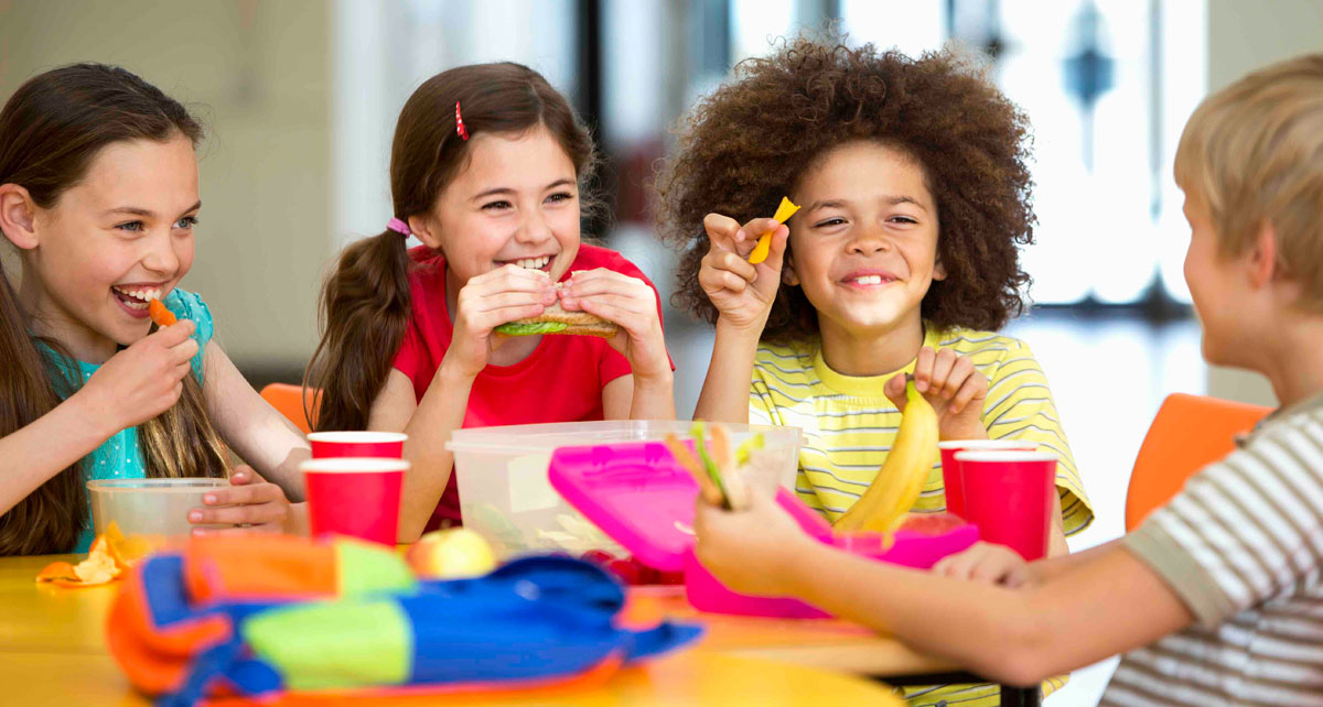 Four children sit around a table, smiling and eating. The table is filled with food containers and drinks, with school bags nearby.