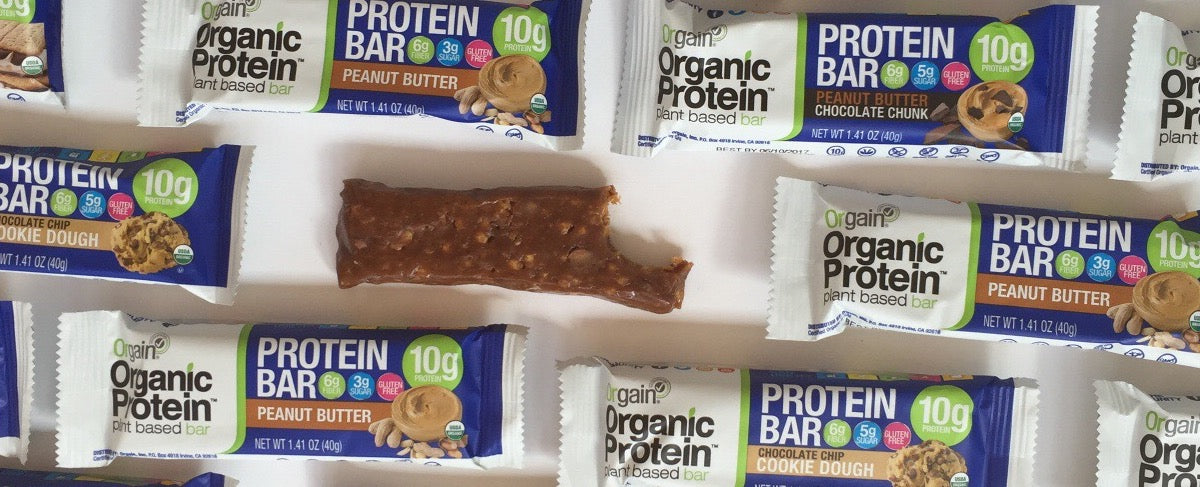 Several Orgain Organic Protein Bars in various flavors such as Peanut Butter, Chocolate Chip Cookie Dough, and Peanut Butter Chocolate Chunk, arranged around one partially eaten bar, with their colorful packaging visible.