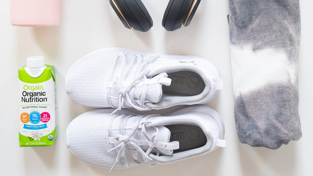 Flat lay of workout essentials including white athletic shoes with laces, a gray and white tie-dye cloth, a pair of black headphones, a carton of Orgain Organic Nutrition drink, and a pink water bottle.