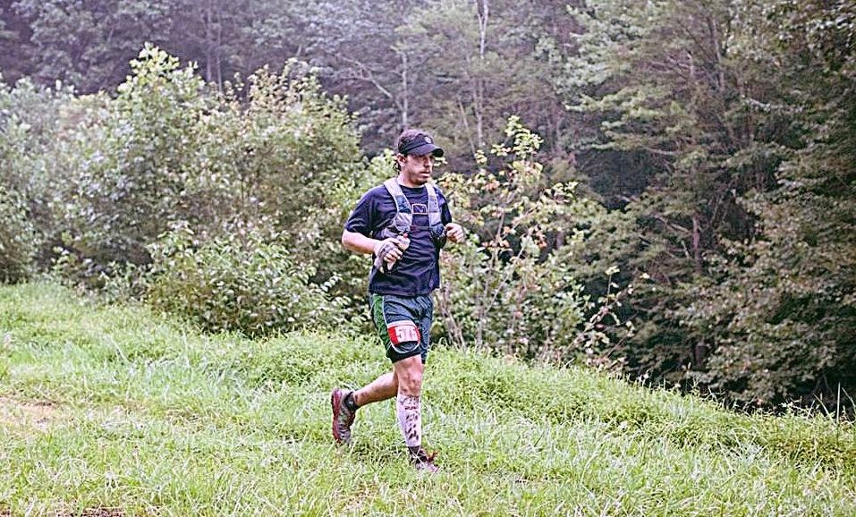 A person wearing athletic gear, a bib number, and a hydration pack runs on a grassy path surrounded by lush greenery and trees. The runner is mid-stride, and the scene appears to be part of a trail in a forested area.