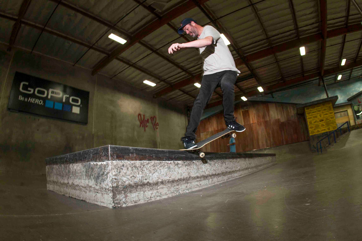 A skateboarder wearing a white shirt and dark pants performs a grind trick on a concrete ledge inside an indoor skatepark. The skatepark has dim lighting and a GoPro advertisement visible in the background.