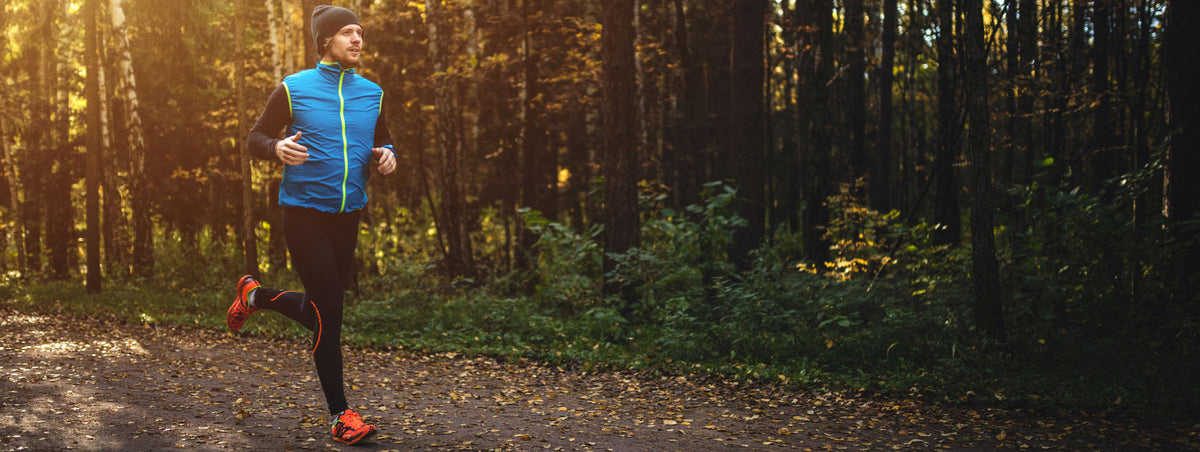 A person wearing a blue vest, black leggings, and red shoes is jogging on a path in a forest during autumn. The sun is shining through the trees, creating a warm and serene atmosphere.