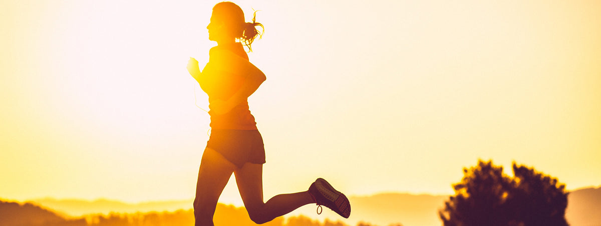 Silhouetted woman jogging at sunrise or sunset, with sunlight creating a glowing effect around her. She is wearing athletic gear, including shorts and a t-shirt. The background features a softly blurred landscape with trees and hills.