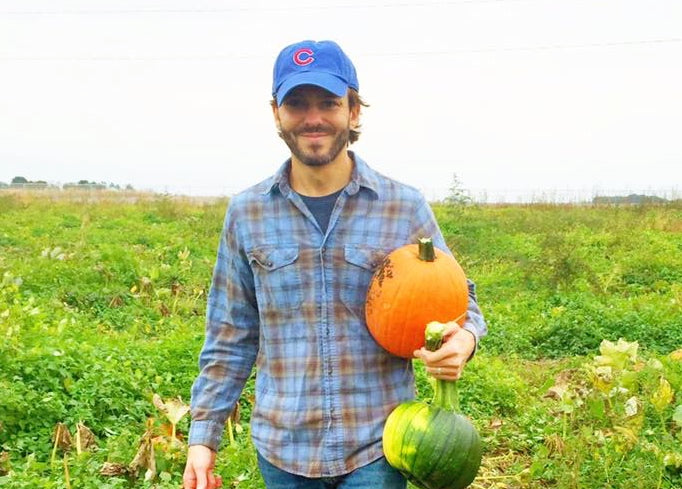 A man in a blue plaid shirt and blue baseball cap stands in a pumpkin patch. He is holding a large orange pumpkin in one hand and two smaller green fruits in the other. The background shows a lush, green field.