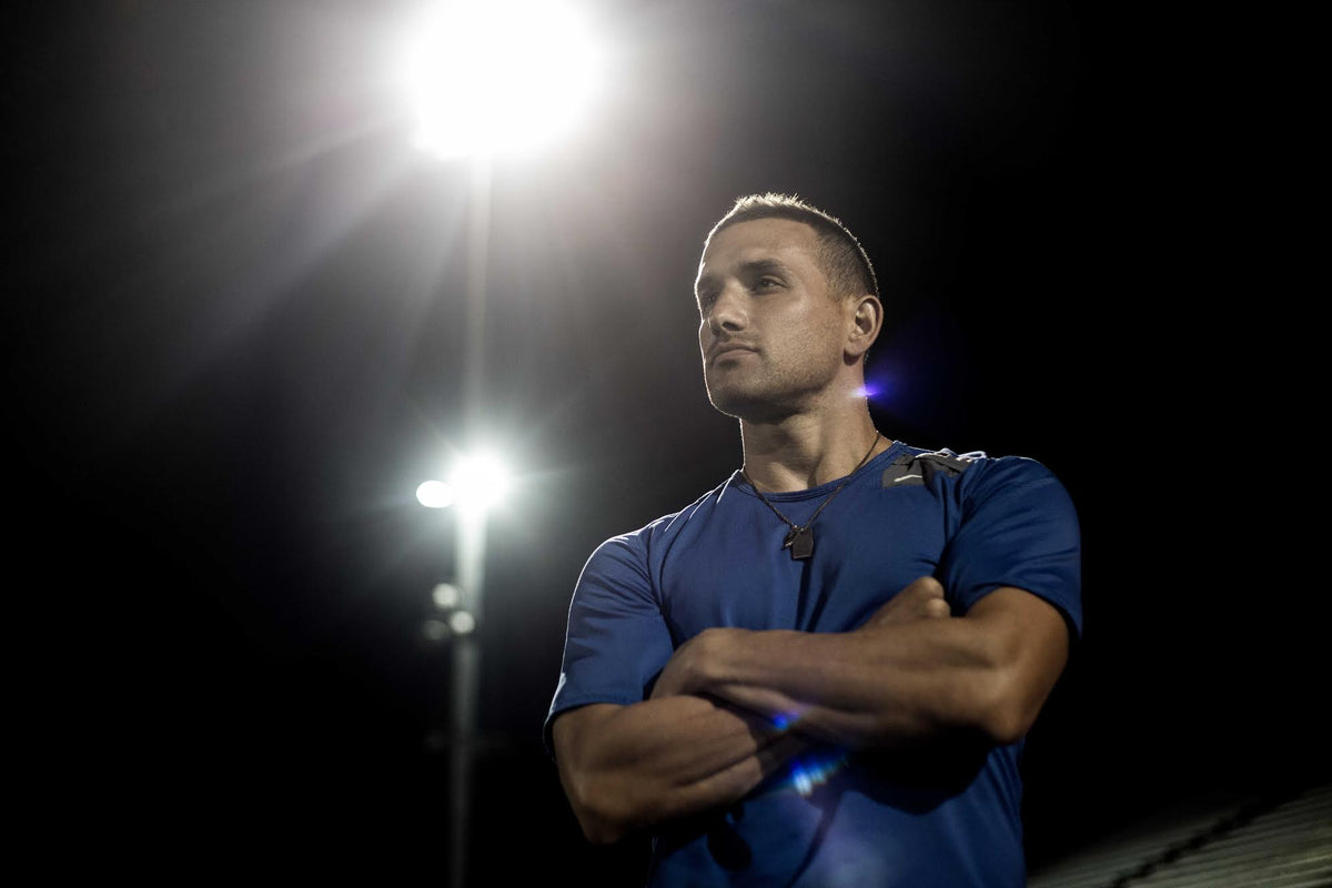 A man in a blue shirt stands with arms crossed under a bright light at night. The light creates a dramatic effect, illuminating his face and casting shadows, evoking a sense of determination.