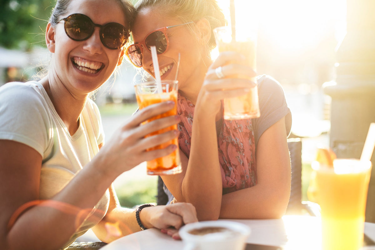 Two smiling women are sitting outside at a cafe, enjoying drinks in tall glasses with straws. They are wearing sunglasses and appear to be in a cheerful mood, with sunlight illuminating the scene.