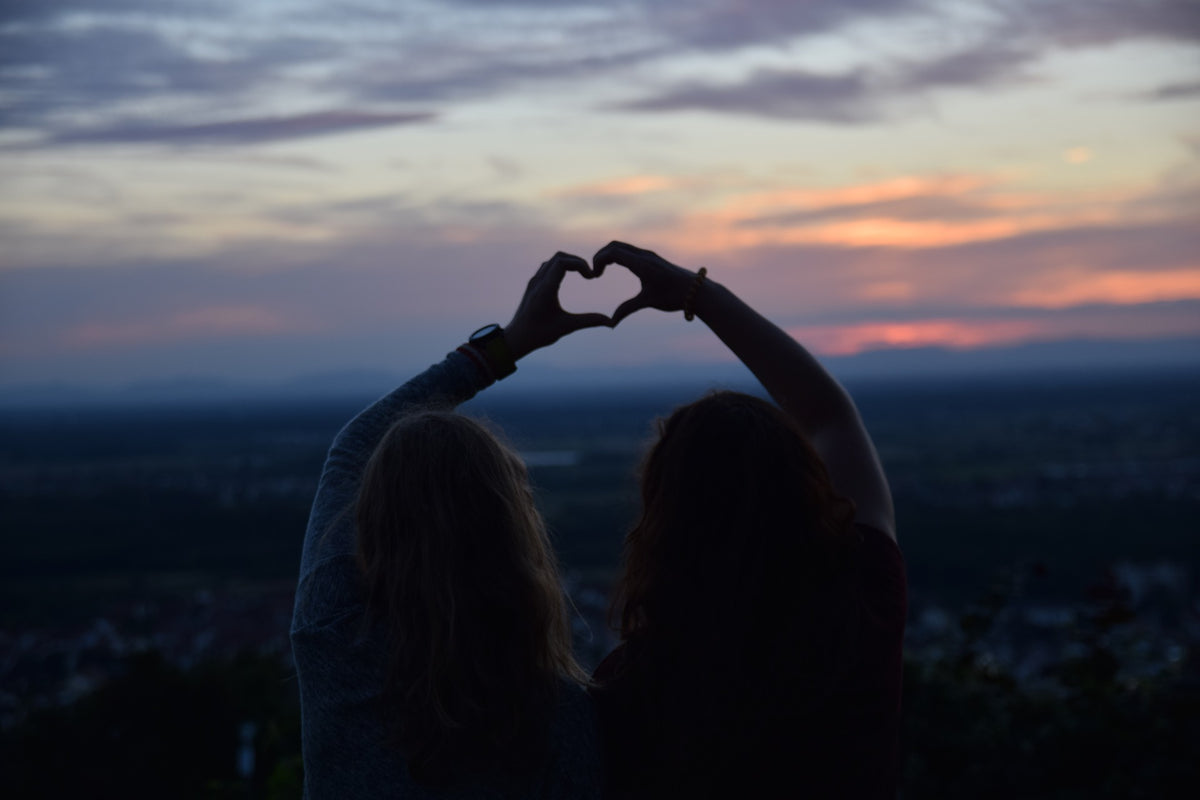 Two women watching the sunset and making a heart with their hands.