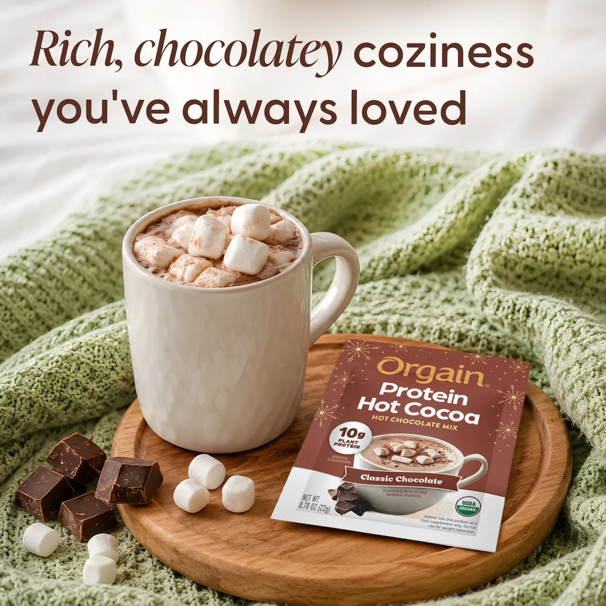 A mug of hot chocolate topped with marshmallows sits on a wooden tray beside an Orgain Protein Hot Cocoa packet, with chocolate pieces and marshmallows scattered around on a green knit blanket. Text at the top reads, 'Rich, chocolatey coziness you've always loved.