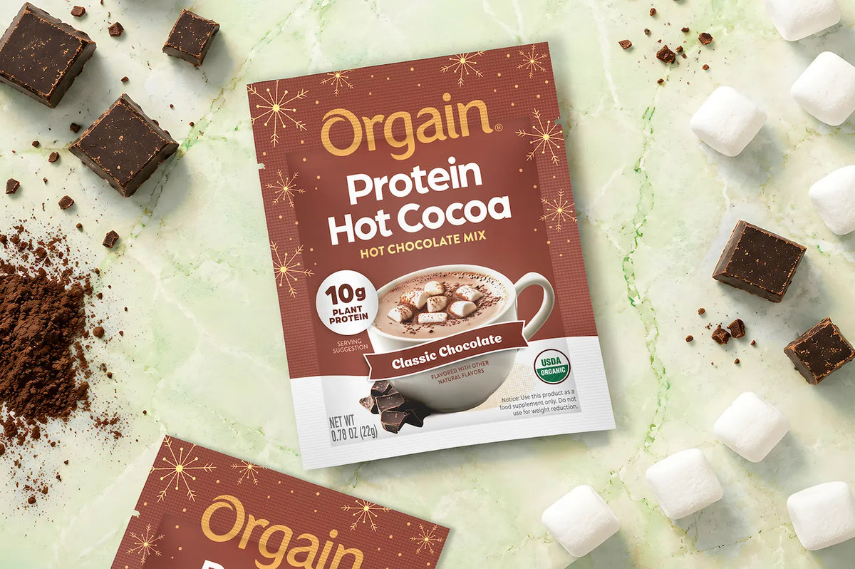 A package of Orgain Protein Hot Cocoa Classic Chocolate Mix is shown on a light marble surface surrounded by chocolate pieces, cocoa powder, and marshmallows.