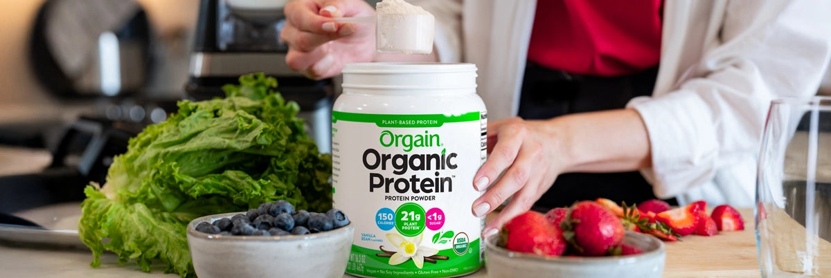 Person holding a container of Orgain Organic Protein with fruits and vegetables on a table.