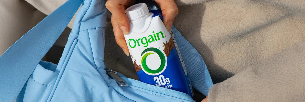 Person holding an Orgain protein shake container with a blue bag in the background