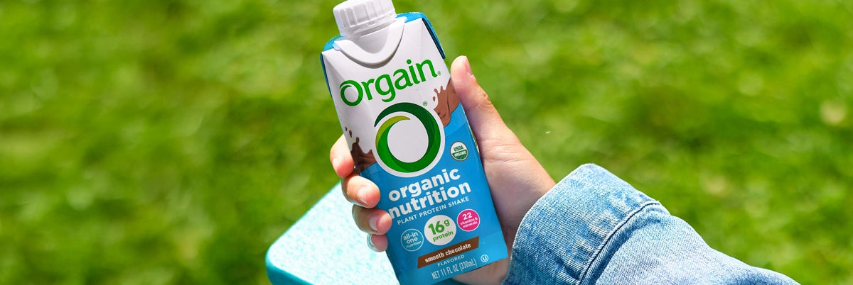 Hand holding a Orgain protein shake bottle against a green outdoor background